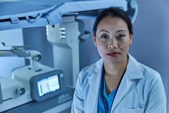 Radiologist looking into camera in front of a Hologic Affirm Prone Biopsy table.